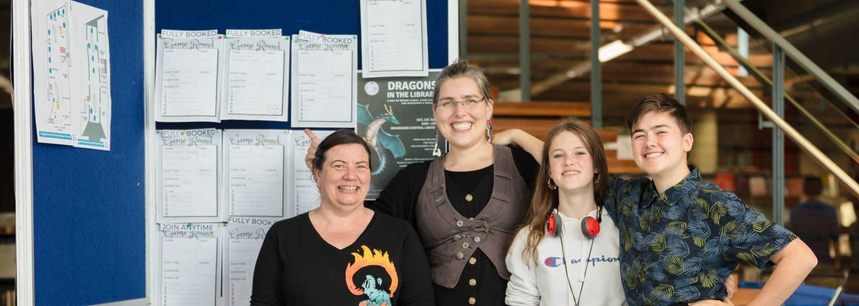 Four smiling people in the library, standing in front of a black board with game descriptions.