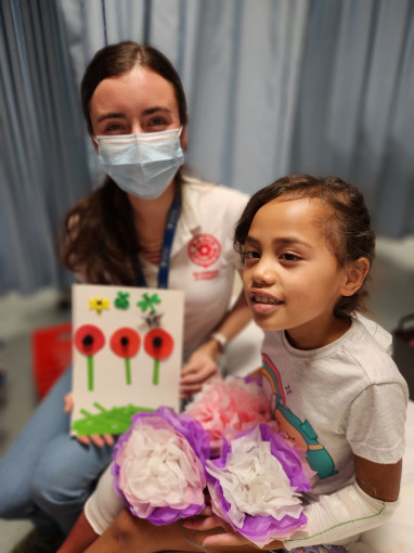 A smiling child in hospital with a volunteer, presenting cards and paper flowers they made together.